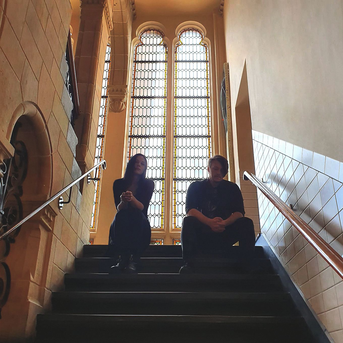 Two people are sitting in a stairwell, silhouetted against  a large church-like window behind them